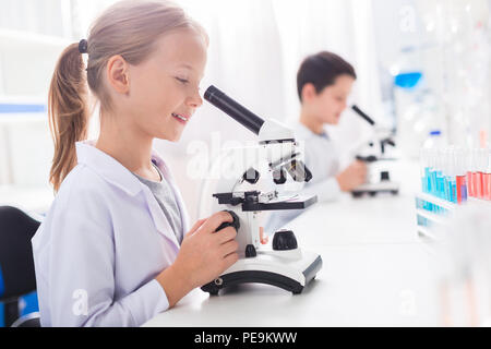 Even closer. Nice smart busy girl learning while using microscope and wearing lab coat Stock Photo