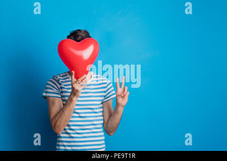 Portrait of a young man in a studio on a blue background, holding red heart. Stock Photo