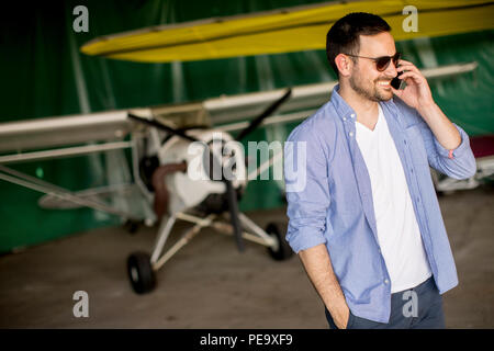 Young handsome young pilot checking airplane in the hangar and using mobile phone Stock Photo