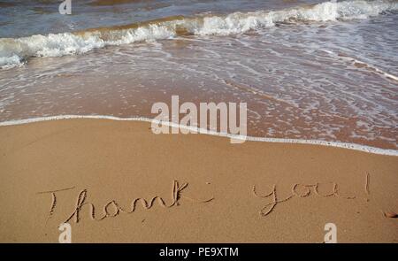 A beautiful gratitude message "Thank you" handwritten in a cursive style on the red sand on a beach with a wave coming in, Prince Edward Island. Stock Photo