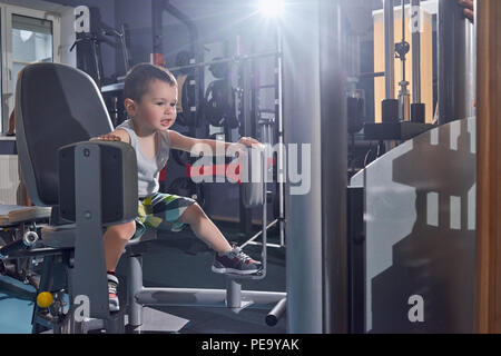 Cute little boy trying to train on heavy metallic simulator in gym. Looking forward, wearing wide shorts, grey tshirt, sport sneackers. Keeping healthy lifestyle from childhood, future sportsman. Stock Photo