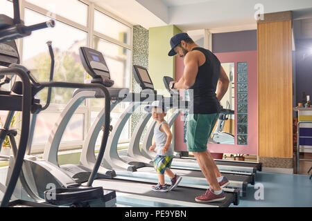 Young father running on racetrack in gym with his little son. Maintaining healthy lifestyle, having strong, muscular body, spending family time together. Little boy wearing wide shorts, tshirt, cap. Stock Photo