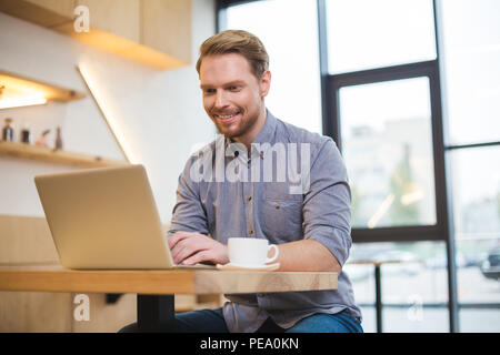 Professional freelancer. Cheerful nice positive delighted man sitting at the table and working on the laptop while being a freelance Stock Photo