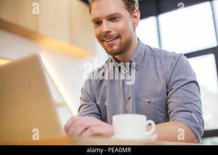 Positive mood. Delighted nice joyful man looking at the laptop screen and smiling while being in the positive mood Stock Photo