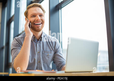 Good mood. Cheerful positive nice man putting his cell phone to the ear and laughing while having a pleasant conversation Stock Photo