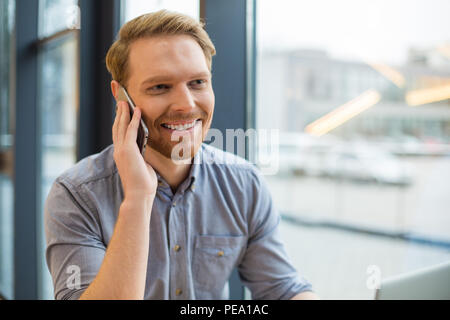 Pleasant communication. Positive nice joyful man smiling and having a pleasant phone conversation while being in the cafe Stock Photo