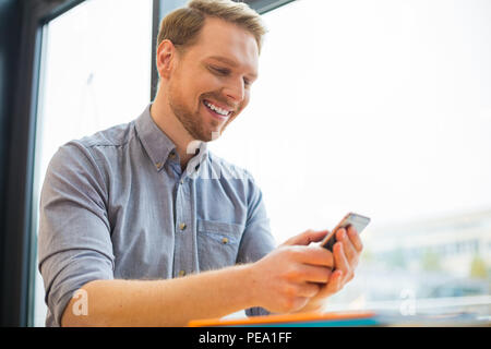 Modern technological device. Cheerful happy delighted man looking at the smartphone screen and smiling while using this device Stock Photo