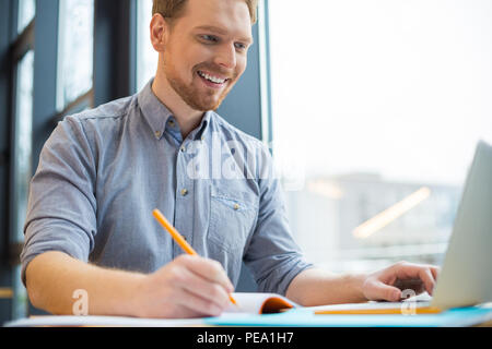 Hard working person. Cheerful nice positive man sitting at the table and taking notes while being involved in his work Stock Photo