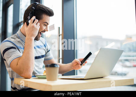 So exiting. Happy nice positive man holding his smartphone and expressing emotions while listening to music in the headphones Stock Photo