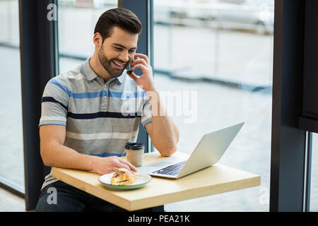 Mobile call. Delighted nice happy man smiling and sitting on front of his laptop while talking on the phone Stock Photo