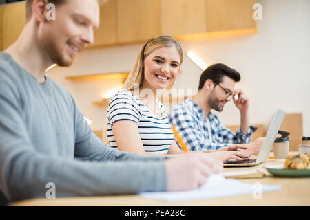 Great mood. Nice positive joyful woman sitting together with her colleagues and looking at you while working on the laptop Stock Photo