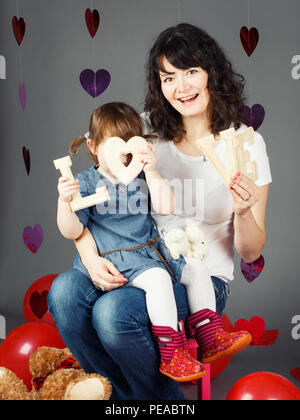 Portrait of young Caucasian mother sitting on chair with baby girl toddler on her laps knees in studio holding wooden letters love smiling laughing  h Stock Photo