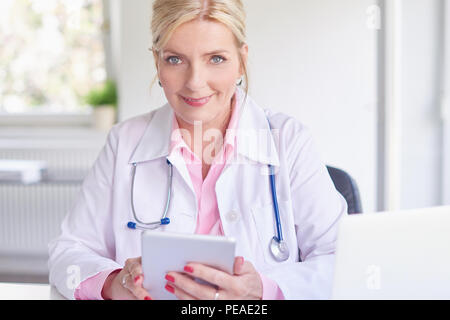 Senior female doctor using touchpad while sitting at doctor's room in front of laptop and computer. Stock Photo