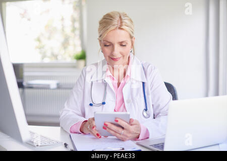 Senior female doctor using touchpad while sitting at doctor's room in front of laptop and computer. Stock Photo