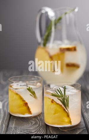 jug of lemonade with pineapple and rosemary, two glasses of lemonade with pineapple pieces, ice cubes and rosemary on grey wooden tabletop Stock Photo