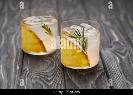 two glasses of lemonade with pineapple pieces, ice cubes and rosemary on grey wooden tabletop Stock Photo