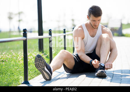 Full length portrait of handsome young man tying sports shoes while sitting on running track in park, copy space Stock Photo