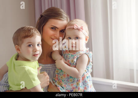 Portrait of beautiful white Caucasian woman mother hugging two cute adorable children kids, son and daughter, boy and girl, sitting on bed near window Stock Photo