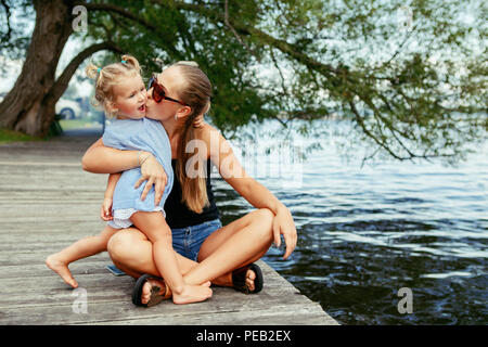 Group portrait of happy white Caucasian mother and daughter child having fun outside. Mom and child girl playing, hugging kissing in park on pier by w Stock Photo
