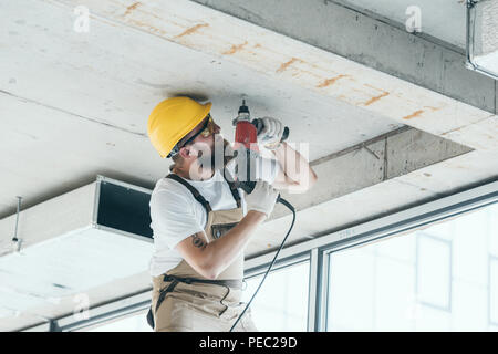 low angle view of builder in protective googles and hardhat working with drill at construction site Stock Photo