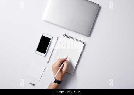 cropped shot of man writing in notebook at workplace with gadgets on white surface for mockup Stock Photo