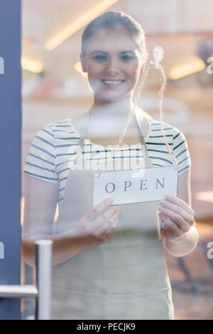 In the morning. Cheerful nice positive woman standing behind the glass door and smiling to you while opening the cafe in the morning Stock Photo
