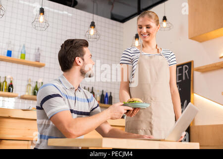 Thank you. Happy positive nice man sitting at the table and taking a plate with hamburger while visiting a cafe Stock Photo