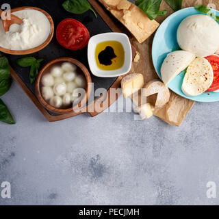 Italian cheeses. Mozzarella Buffalo, crottin, balls, Stracciatella, basil leaves, tomatoes, olive oil with vinegar on gray background. Top view, copy space. Stock Photo