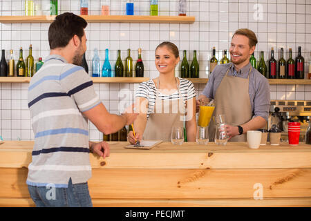 What would you like. Positive delighted young woman smiling and looking at her customer while taking an order from him Stock Photo