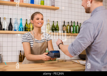 Credit card payment. Joyful nice attractive woman smiling and using a card terminal while taking payment from the customer Stock Photo
