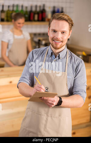 What would you like. Nice positive young man wearing apron and taking notes while listening to the customers order Stock Photo