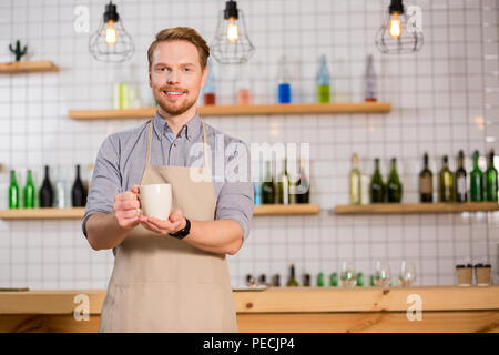 Special drink. Joyful nice handsome man wearing apron and holding a cup of tea while offering it to you Stock Photo