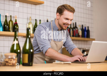 Online order. Delighted nice positive man standing at the counter and typing while working on the laptop Stock Photo