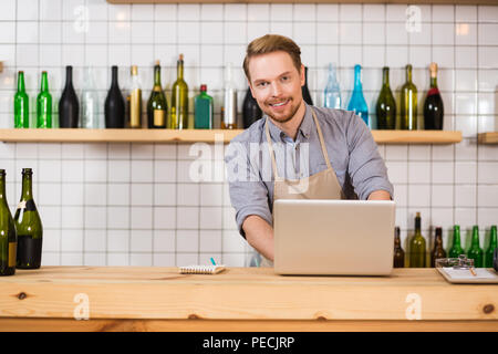 What is your order. Joyful nice pleasant man standing at the laptop and looking at you while taking an order in the cafe Stock Photo