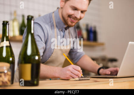 Making a list. Joyful nice pleasant man using a pencil and writing a list while standing at the counter Stock Photo