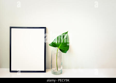 Black photo frame and green plant in a chrystal vase arranged against emty grey wall. Frame mock-up. Stock Photo