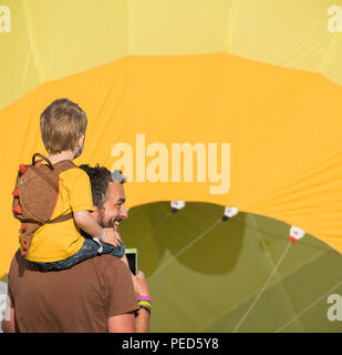 Boy on his dad's shoulders watching a yellow balloon being inflated at the Bristol International Balloon Fiesta. Stock Photo