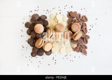 A selection of caramel coloured macarons (macaroons) scattered on a bed of dark, milk and white chocolate buttons and chocolate pieces Stock Photo