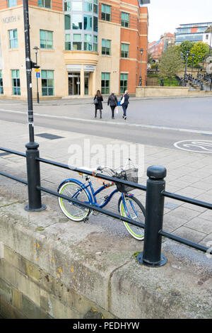 Blue woman's or lady's bike with handlebar basket leaning on and locked to railings on Newcastle Quayside, with three women walking in the background. Stock Photo