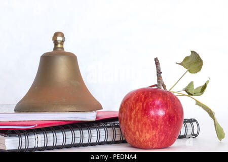 Symbols for education, red delicious homegrown apple with green leaves, brass school bell stacked on black spiral bound sketch notebook, red folder an Stock Photo