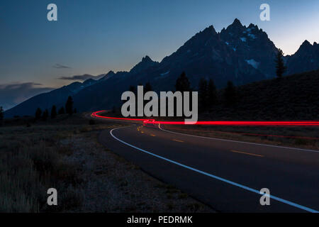 Tail lights in the night highway at Grand Teton National Park with background of mountain range and sun setting behind. Stock Photo