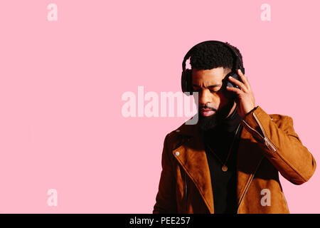 Portrait of a cool man with beard touching his headphones listening to music, isolated on pink studio background Stock Photo