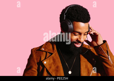 Portrait of a smiling man with beard and headphones, looking down listening to music, isolated on pink studio background Stock Photo