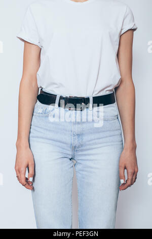 Unrecognizable young woman wearing blanc white t-shirt and blue high waist mom's jeans with belt standing over white background, close-up. Stock Photo