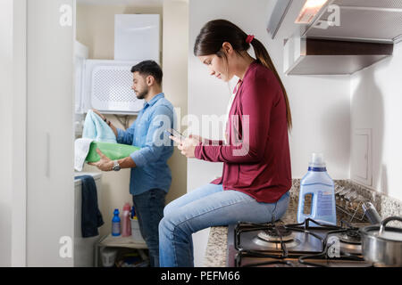 Young couple doing housework. Man putting clothes inside washing machine and girlfriend chatting on mobile phone. Stock Photo