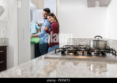 Young couple doing housework. Man putting clothes inside washing machine and girlfriend hugging him. Stock Photo