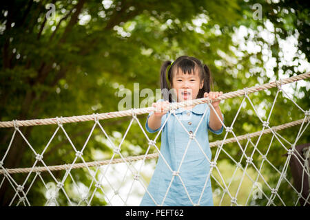 Happy little girl playing climbing on the rope bridge in adventure park colorful garden on hot summer day. Summer activities for kids. Stock Photo