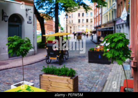 Trishaw rides along the pavement past the outdoor cafe with green plants and flowers in the old center of Riga. Blurry Stock Photo