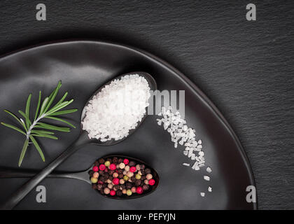 Salt and mixed pepper in dark spoons on black plate on dark slate stone background Stock Photo