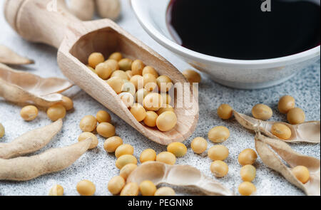 Soybeans in wooden spoon, pods and soy sauce in a white bowl on a gray concrete surface. Selective focus Stock Photo